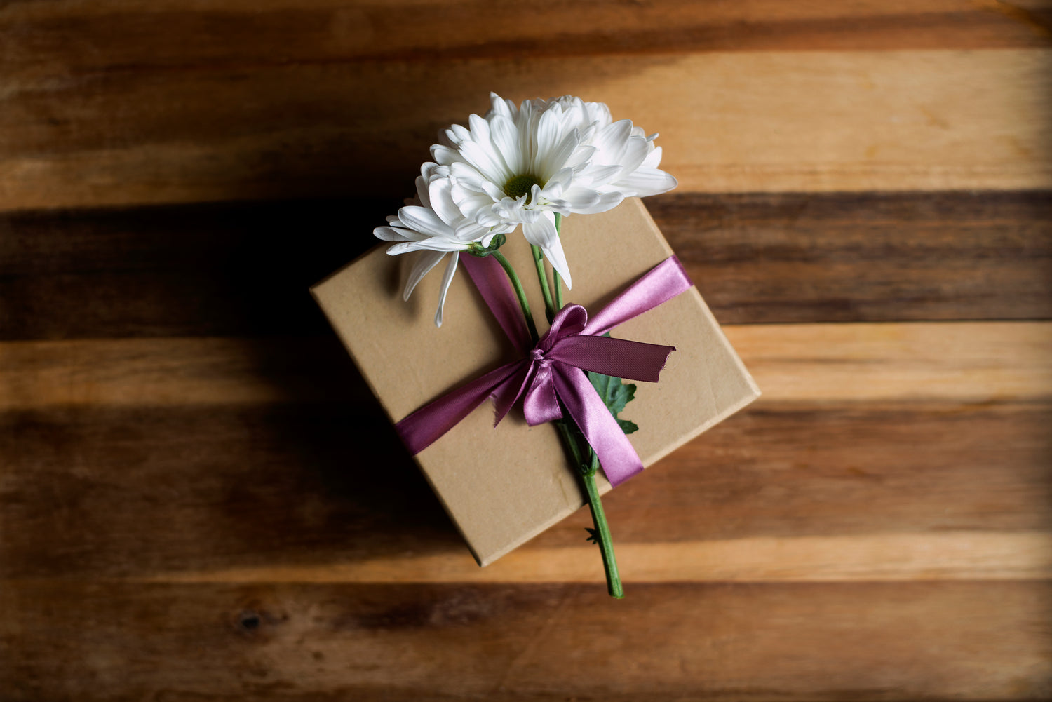 A plain brown box, with a purple ribbon that sits on top of a wooden table. On top of the present is a white flower.